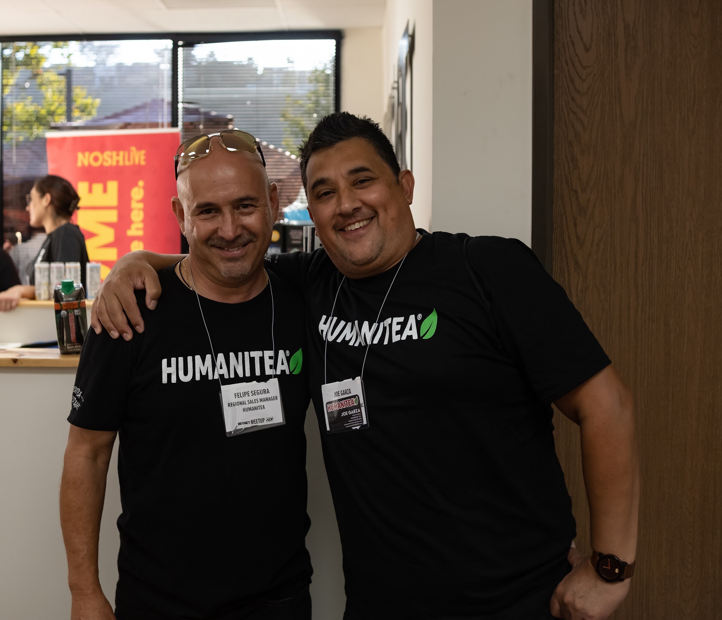 Two smiling men in black Humanitea t-shirts and lanyards stand with their arms around each other in front of a red Nosh Live banner.