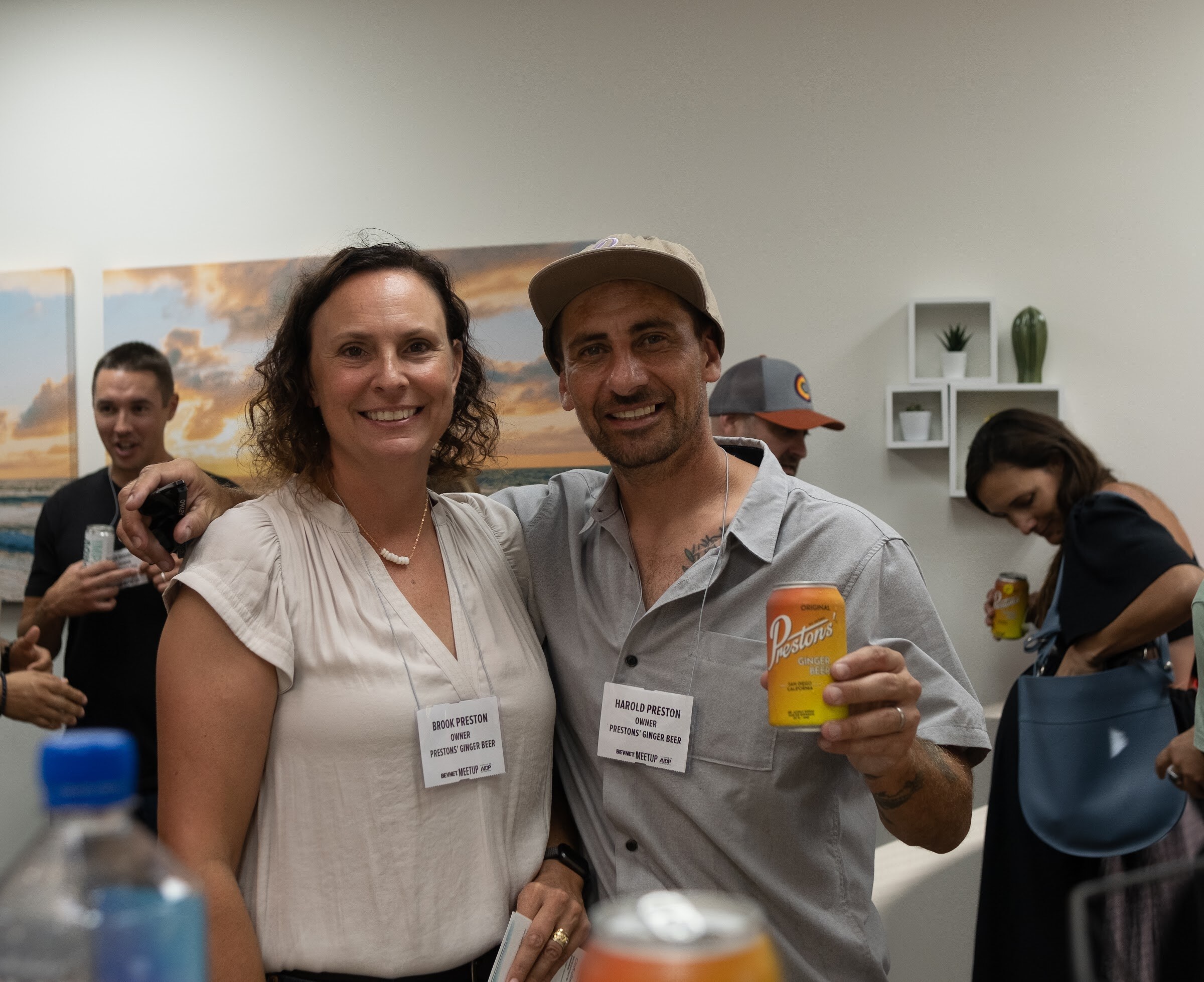 A smiling woman in a cream blouse and a man in a tan cap and gray button-up shirt, both with badges, pose together while he holds up an orange can of Preston's, in front of a wall with a sunset beach photo.