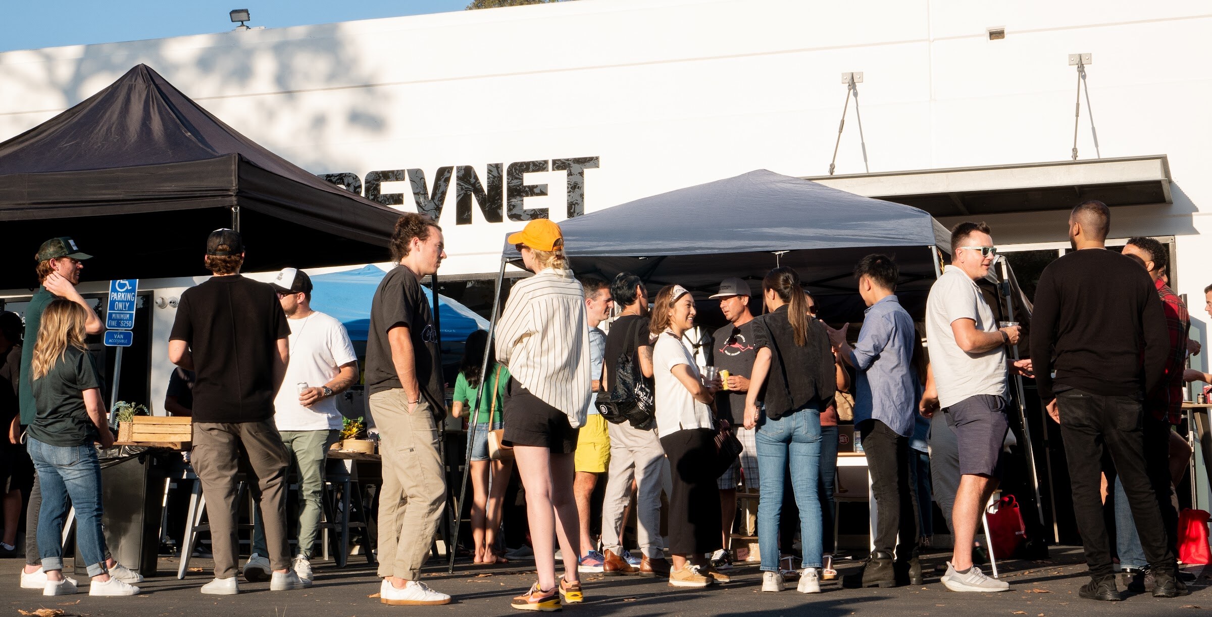A crowd of guests stands in the late-afternoon sun in a parking lot beneath dark and blue canopies in front of a white building with a BEVNET sign.