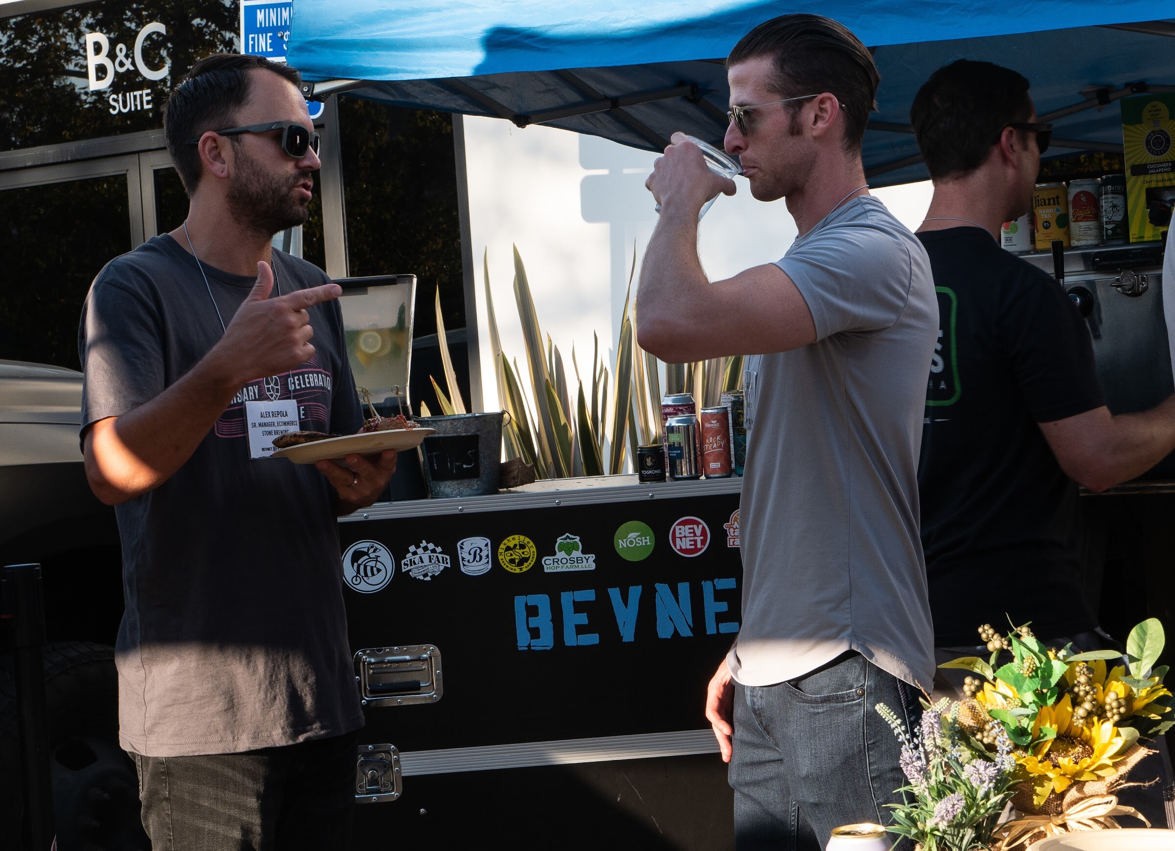 A man in sunglasses and a dark t-shirt holding a plate points while a man in a gray t-shirt drinks from a clear cup, in front of a black road case with BEVNET lettering and brand stickers, under a blue canopy.