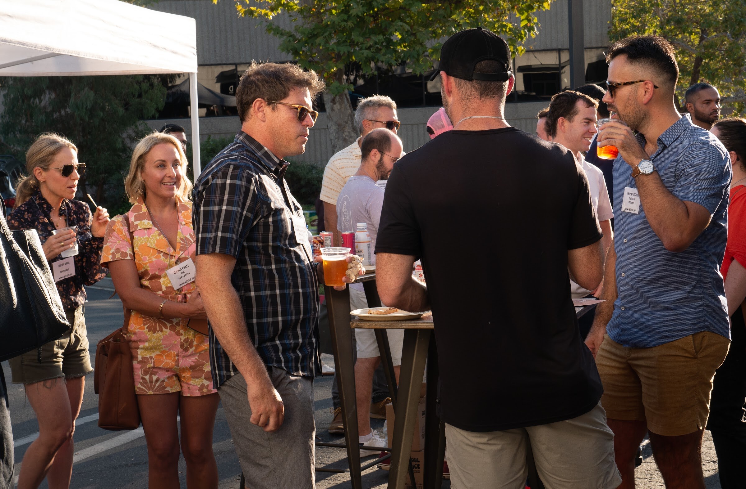A group of guests with name badges talk around a high-top table in the parking lot, including a woman in a floral romper, a man in a plaid shirt, a man in a black t-shirt and cap and a man in a blue shirt drinking a beer, with trees behind them.