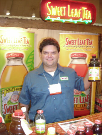 A smiling man in a blue work shirt and badge holds up a bottle of Sweet Leaf Tea. Sweet Leaf Tea banners and more bottles are behind and in front of him, under a neon Sweet Leaf Tea sign.
