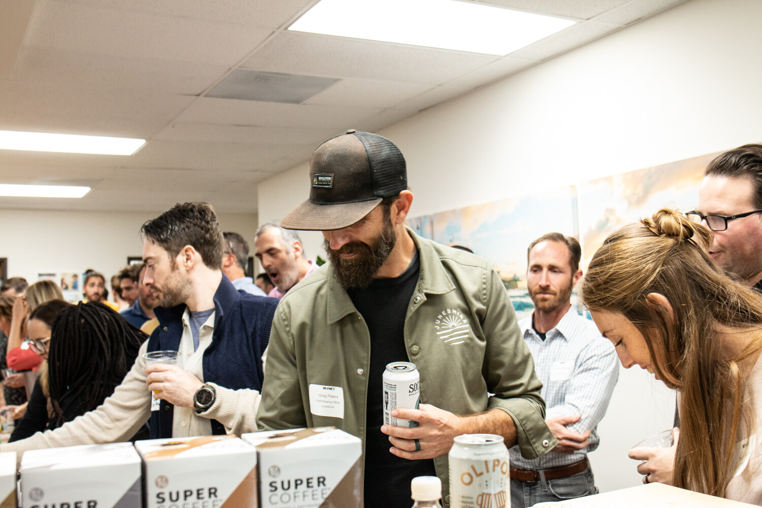 A bearded man in a green jacket holds a can next to Super Coffee boxes while other guests look at products