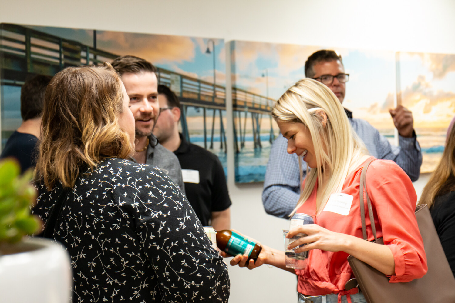 A woman in a coral top looks down at a bottle while others talk in front of a pier mural