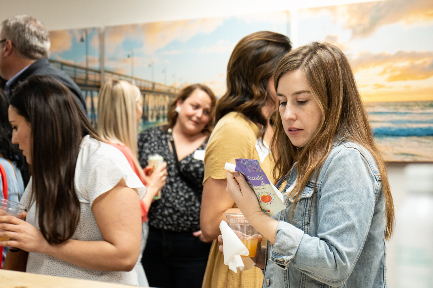 Women holding a drink carton and a cup stand in front of a pier mural with other guests behind them