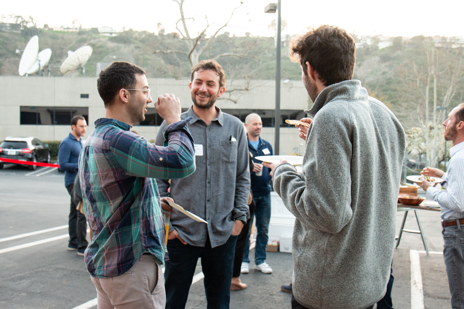 Three men talk outdoors with plates in hand, with a hillside and satellite dishes behind them