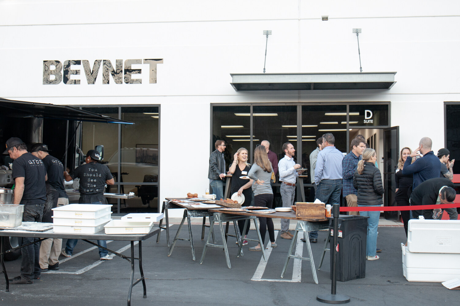 People mingle in front of a white building with BEVNET lettering and a Suite D sign, with catering tables and a rope line in the foreground