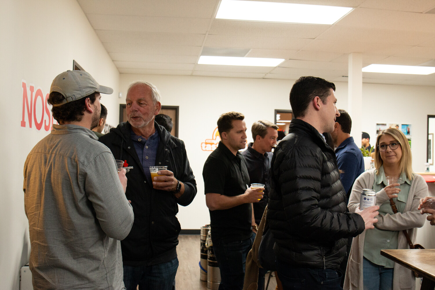 A group of men holding drinks stand in an office hallway with NOSH and BEVNET lettering on the wall behind them