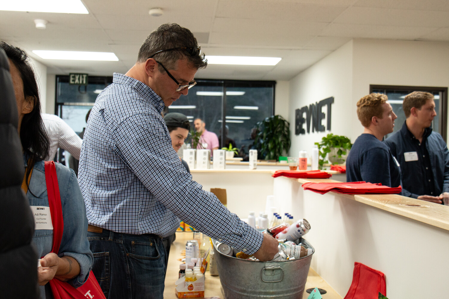 A man in a checked shirt picks a drink from a galvanized tub on the reception counter with a BEVNET sign on the wall behind