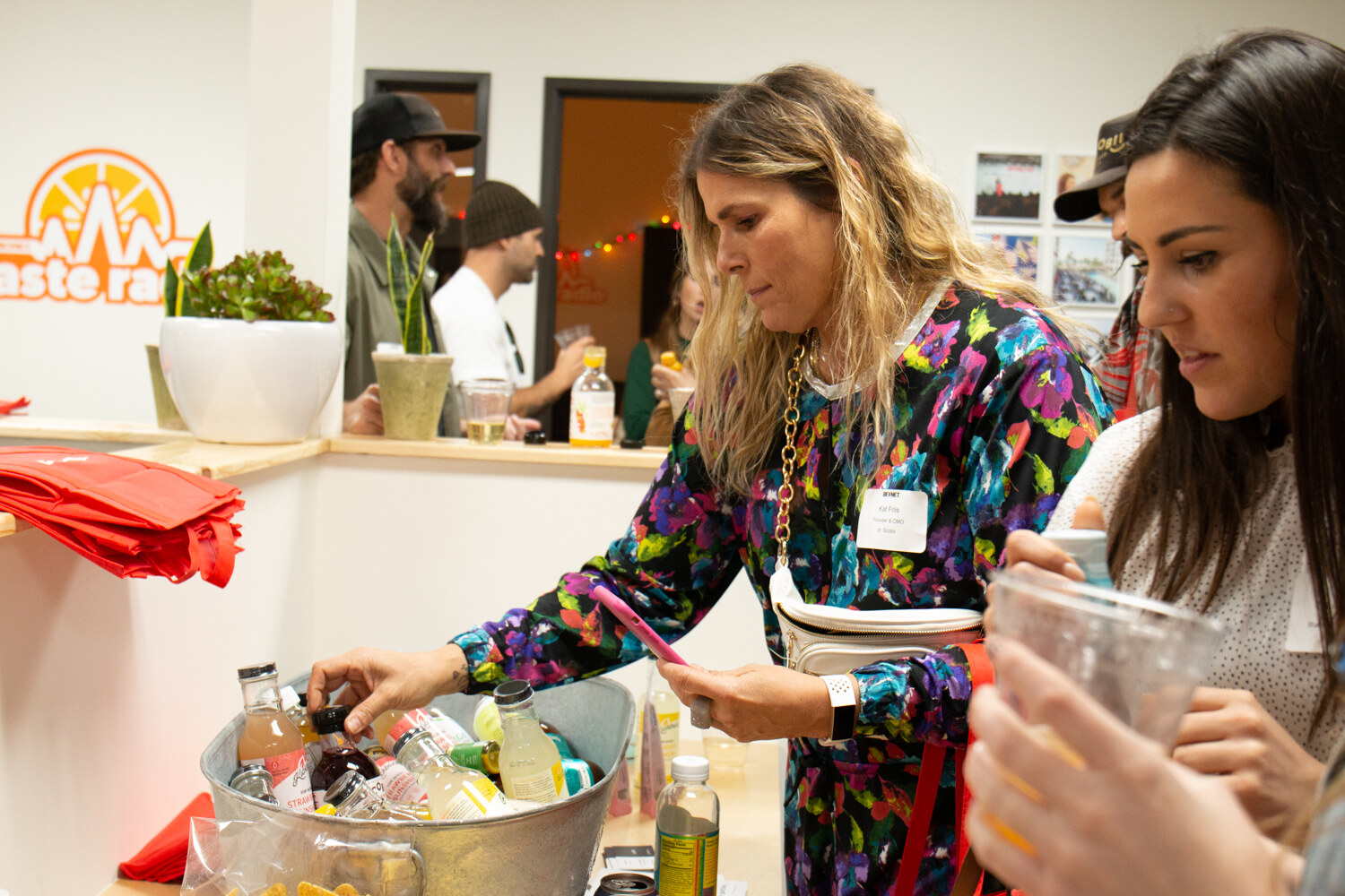A woman in a colorful jacket takes a bottle from a metal tub, with a Taste Radio logo on the wall behind her