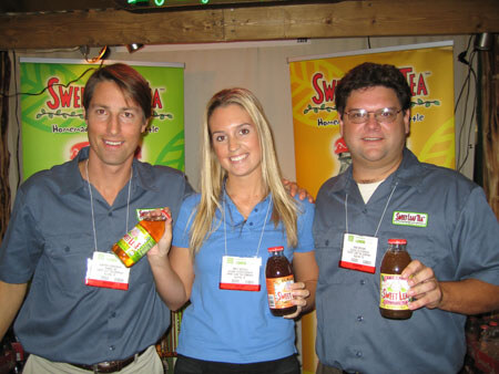 Two men and a blonde woman wearing name badges pose together in front of Sweet Leaf Tea banners, each holding or standing near bottles of tea. One man and the woman are in blue shirts.