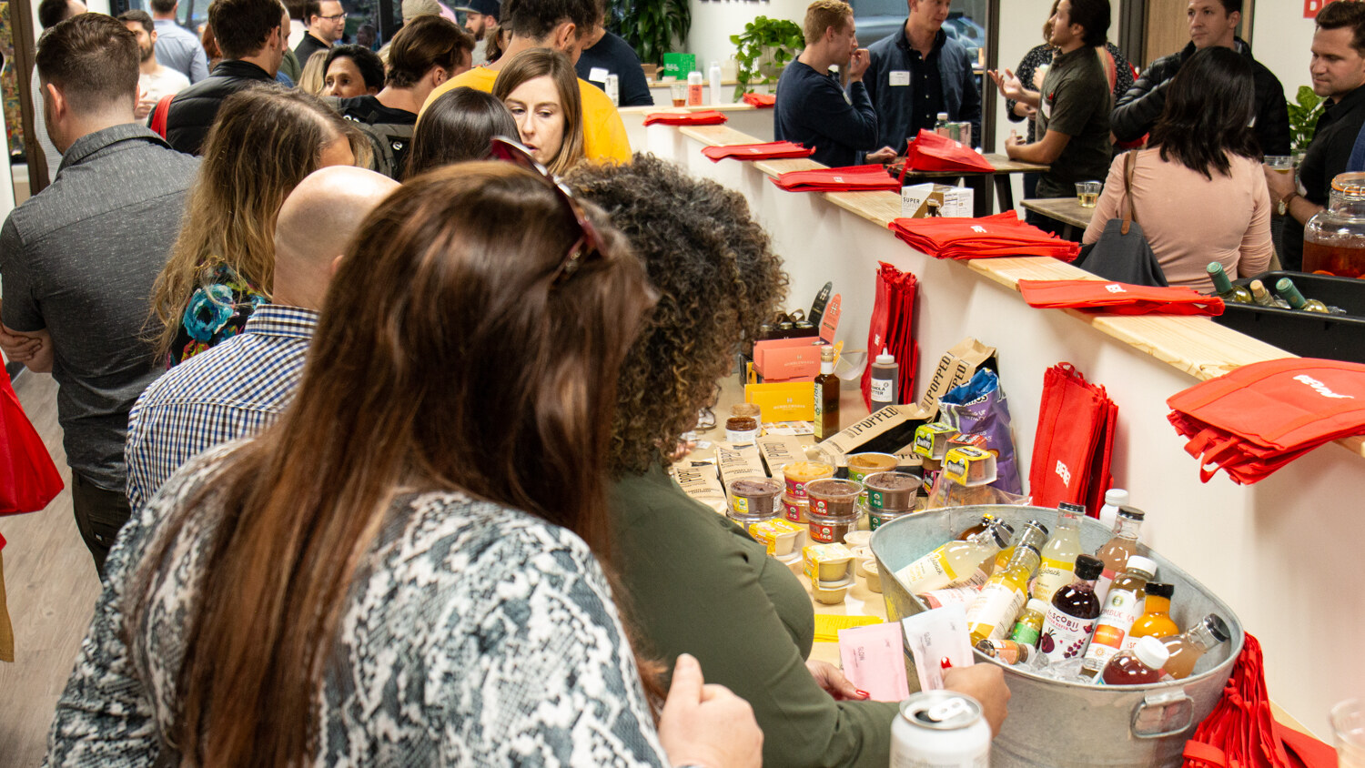 A packed counter of drinks and snacks in an ice tub with guests crowding around and red tote bags stacked behind