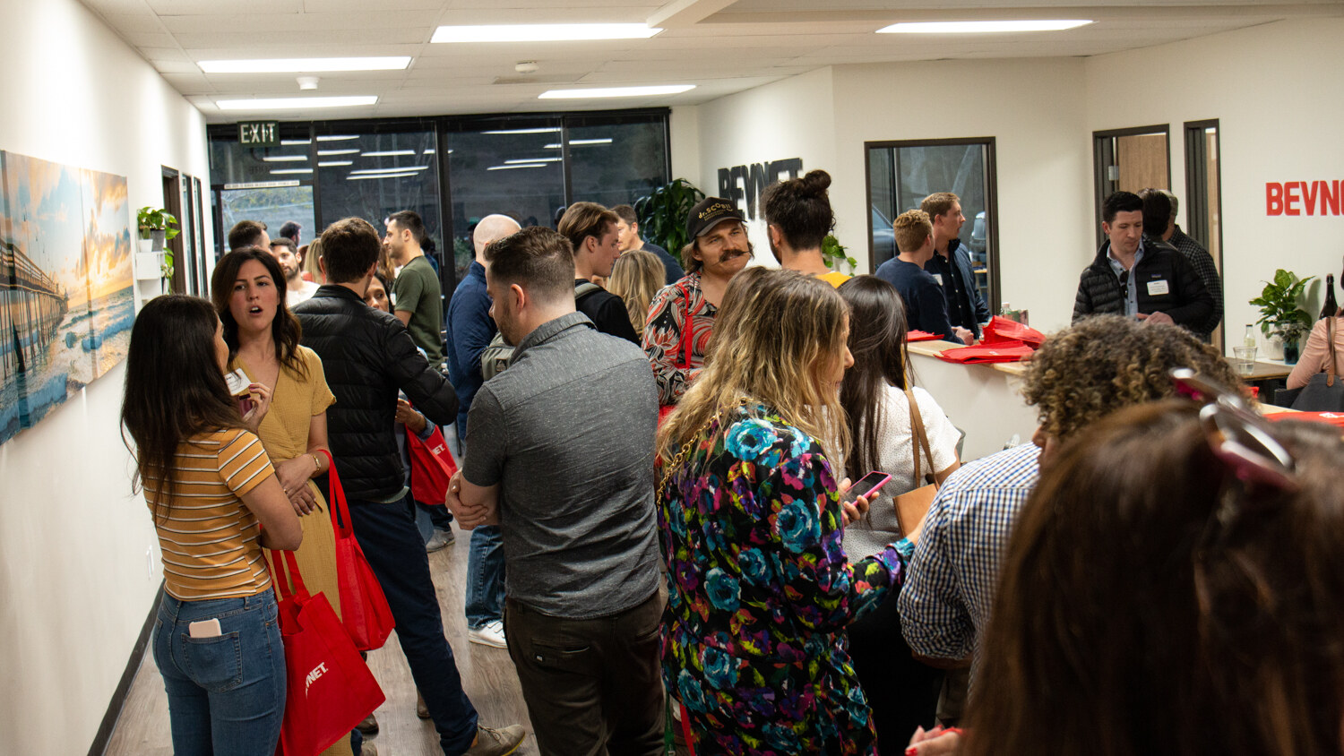 A crowded office hallway with guests, red tote bags and BEVNET lettering on the wall in the background