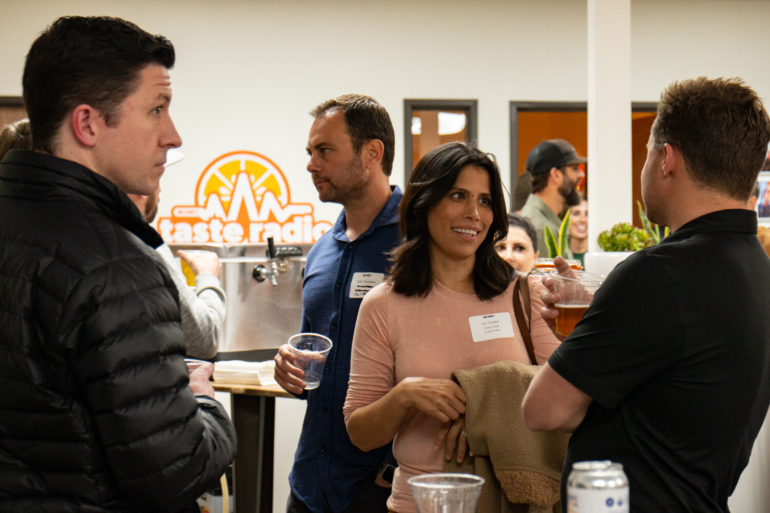 Several guests with drinks and name tags stand in front of an orange Taste Radio logo on a wall