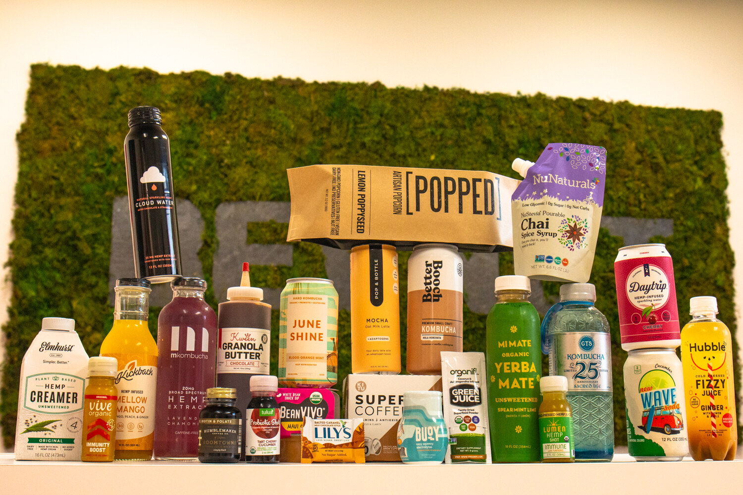A tall arrangement of beverages and snack packages in front of a green moss wall spelling BEVNET, including Hubble, June Shine, Better Booch, Wave and Daytrip