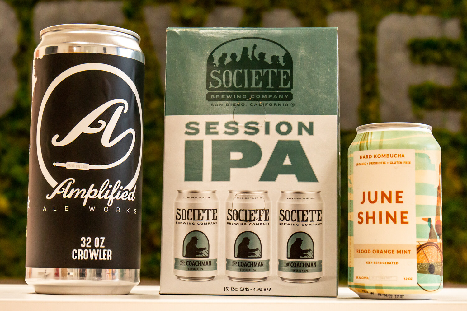 A black Amplified Ale Works crowler, a Societe Brewing Session IPA six-pack and a June Shine can in front of a moss wall
