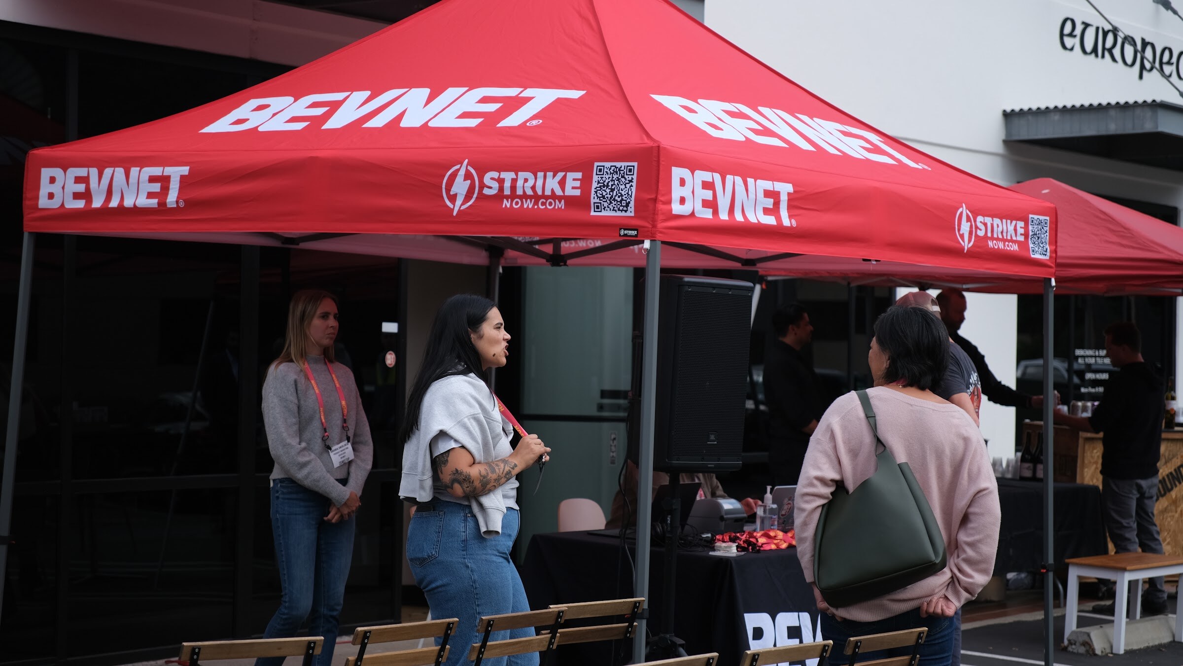 Two women with lanyards stand under a red BEVNET canopy in a parking lot as a third guest walks past.