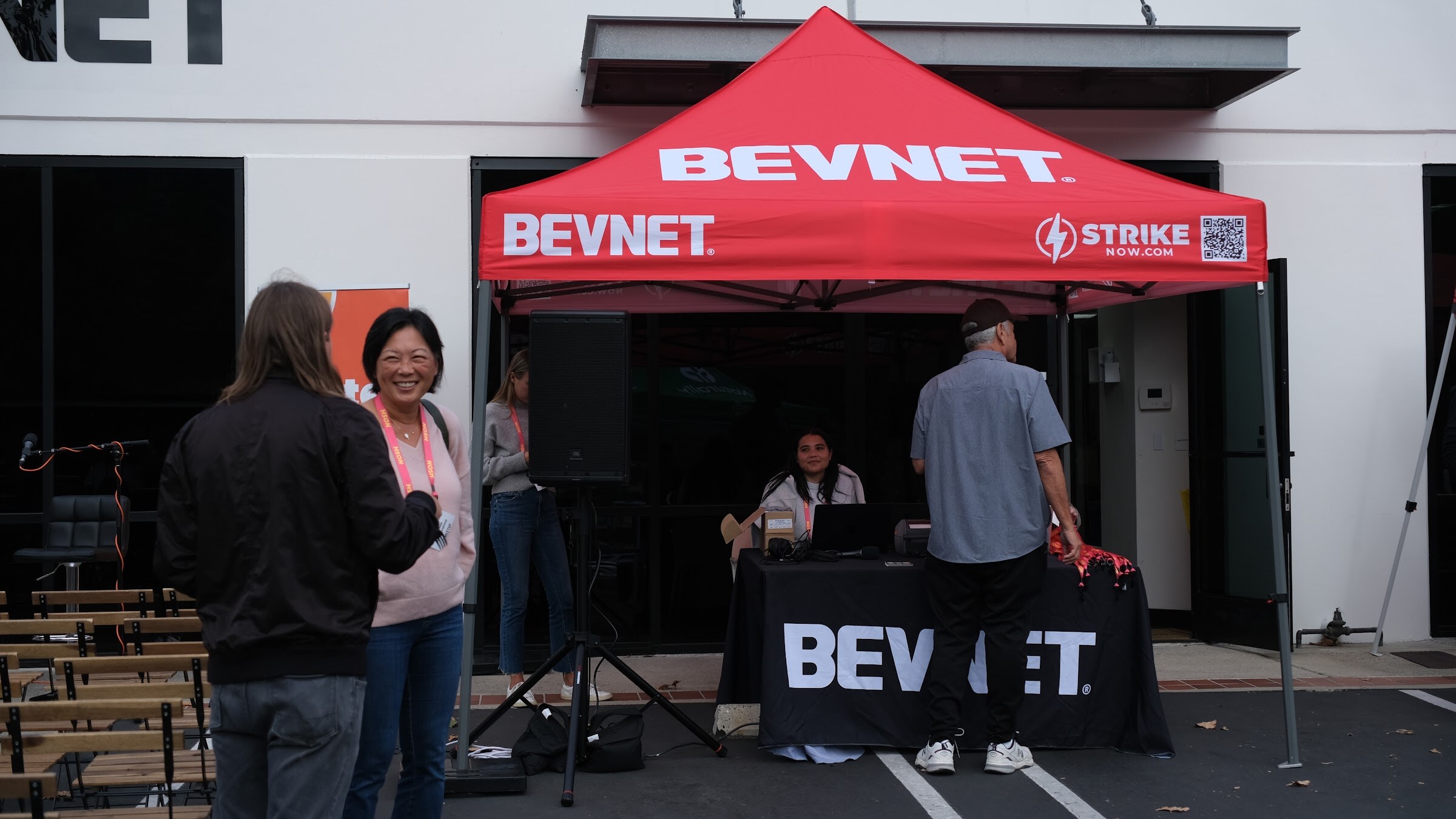 A red BEVNET tent in a parking lot, with a staff member seated at a table behind a black BEVNET banner and two guests talking in front.