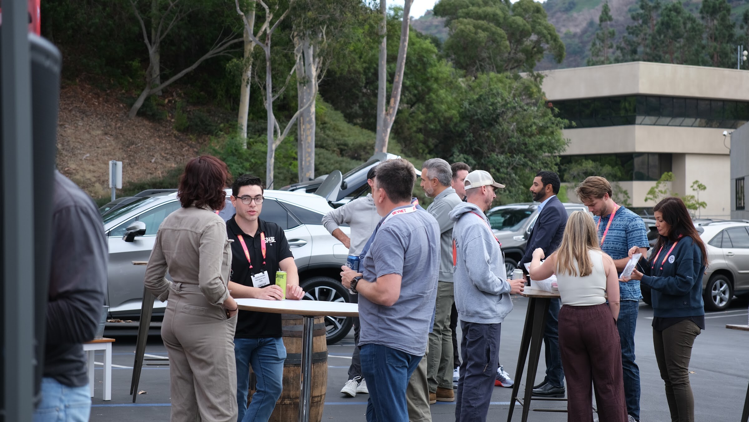 Attendees in lanyards stand around tall tables in a parking lot with eucalyptus trees and hills behind them.