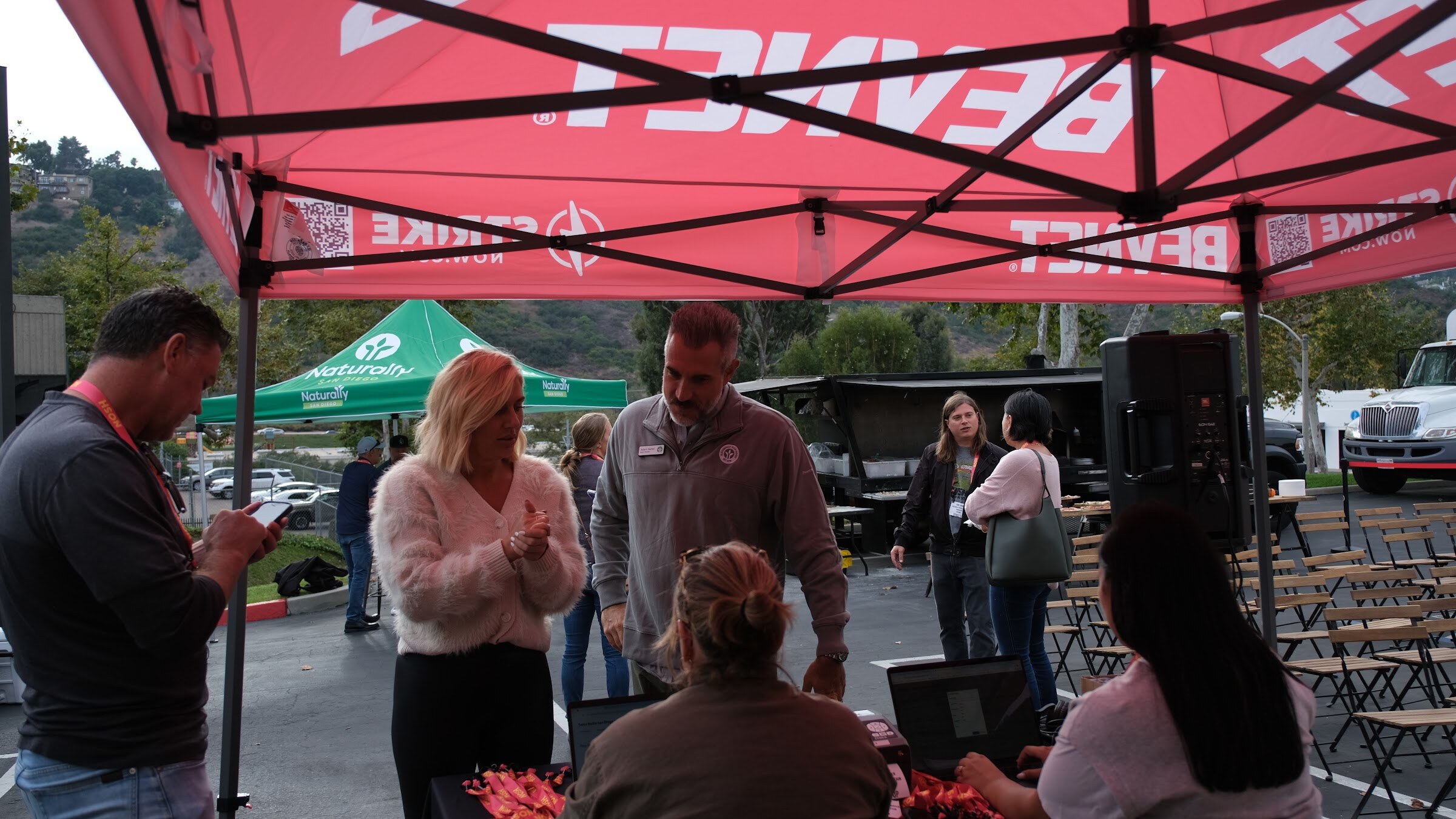 Staff members sit with laptops at a table under a red BevNET canopy while guests in lanyards stand in front.