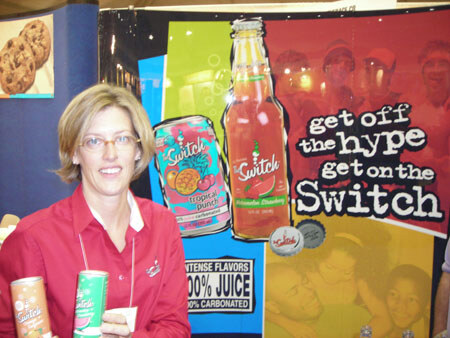 A woman in a red shirt and glasses holds two cans of Switch juice drink in front of a Switch banner reading get off the hype, get on the Switch.