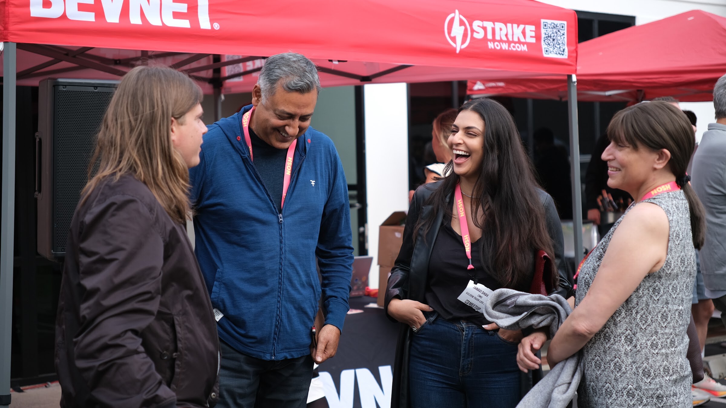 Four attendees wearing lanyards laugh together in front of a red BevNET canopy.