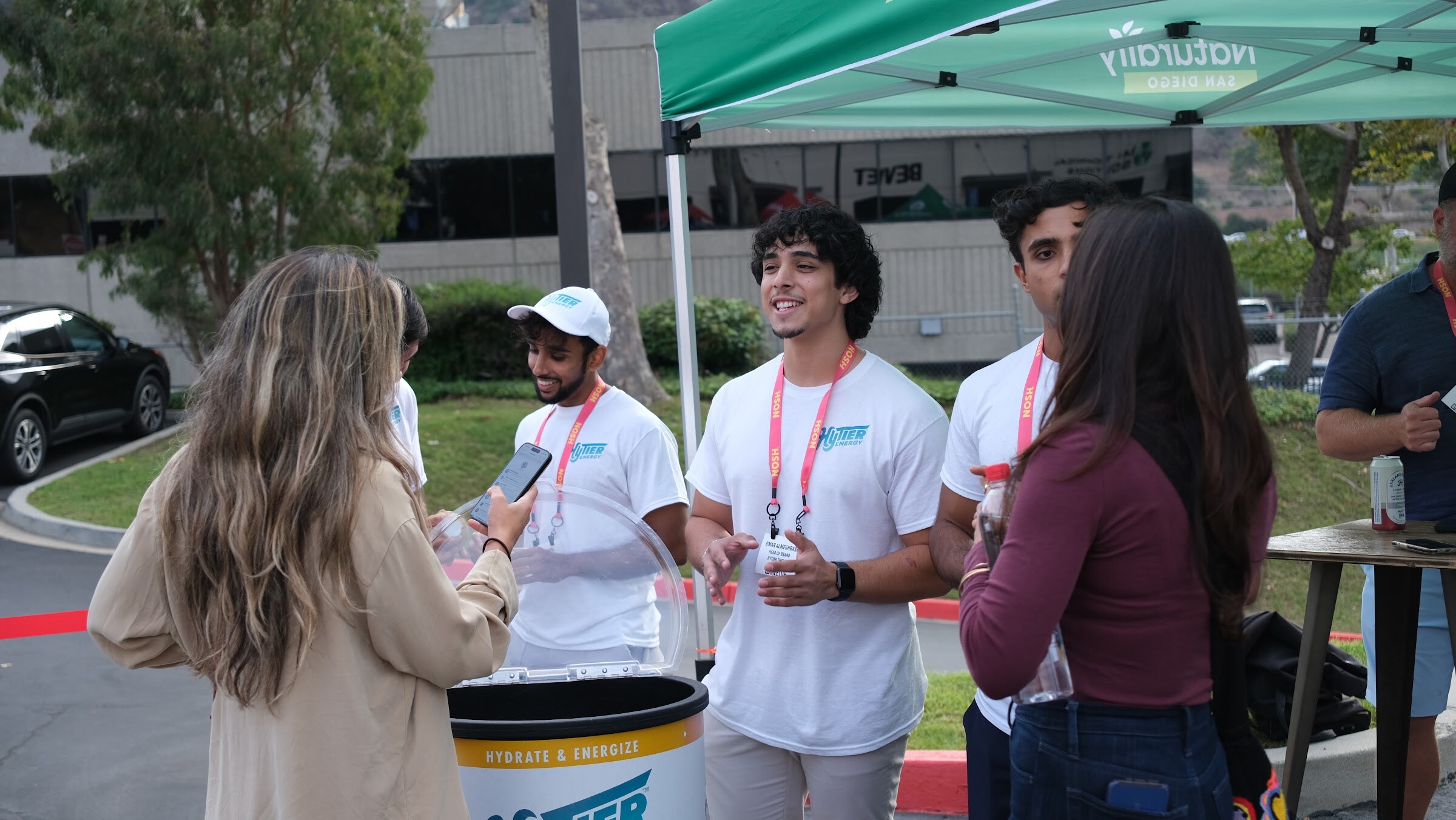 Three men in white T-shirts and lanyards talk with a woman holding a drink beside a cooler under a green tent.