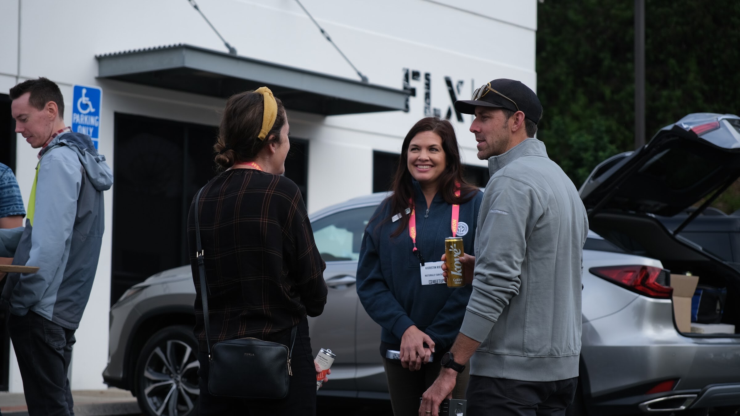 A smiling woman with a lanyard holds a can while talking with three other guests beside a car with an open trunk.