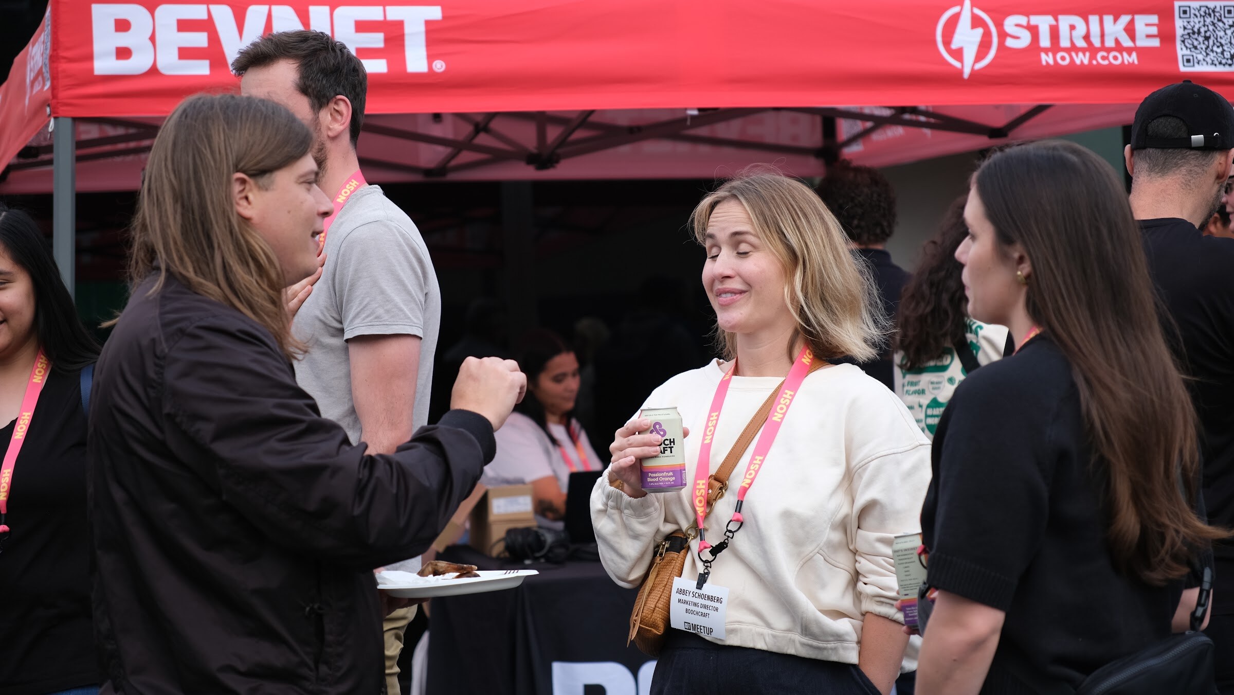 Several people with lanyards talk and laugh under a red BEVNET canopy, one holding a drink and a plate of food.