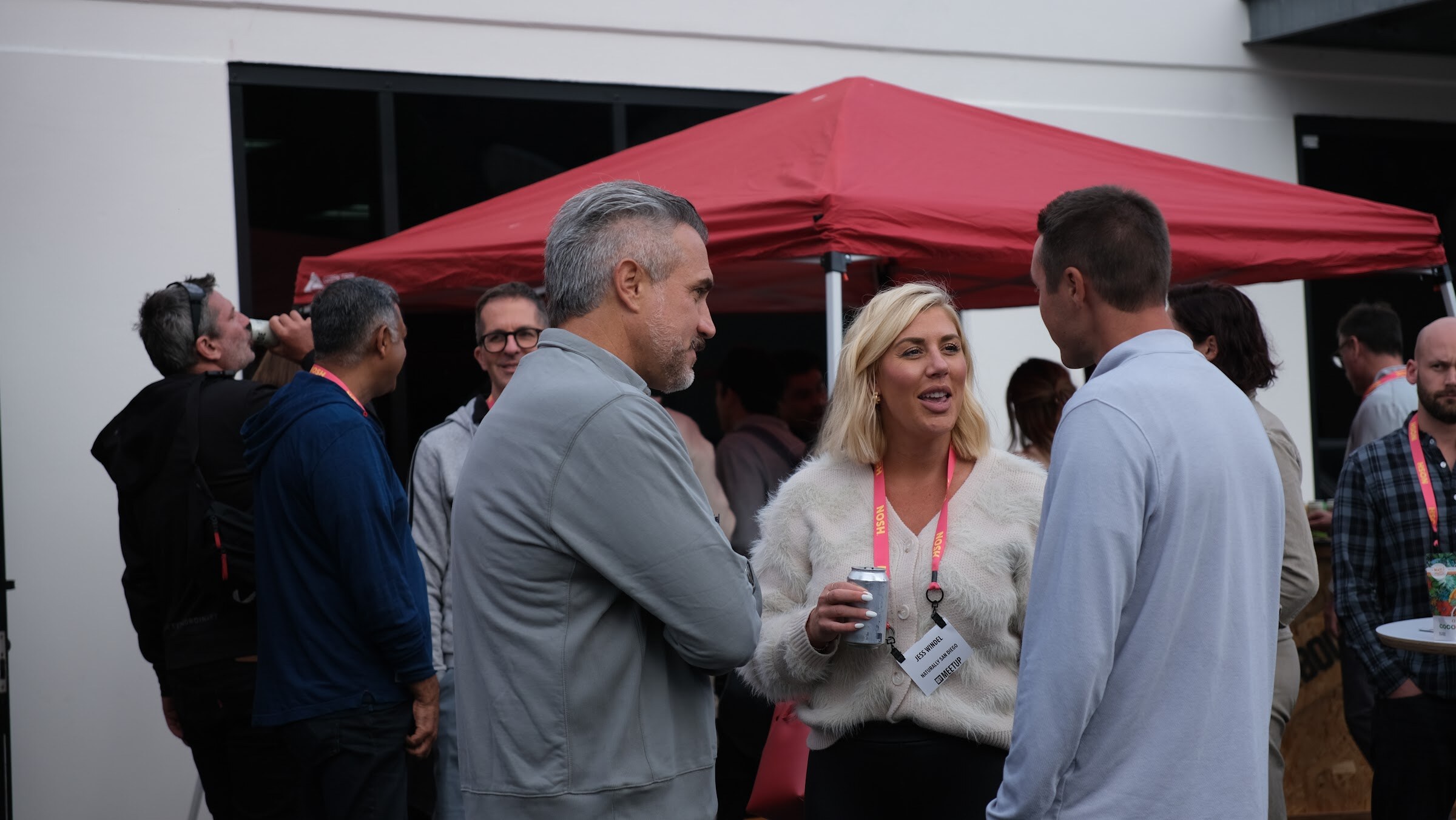 Four attendees with lanyards talk in a group in front of a red tent, one drinking from a can.