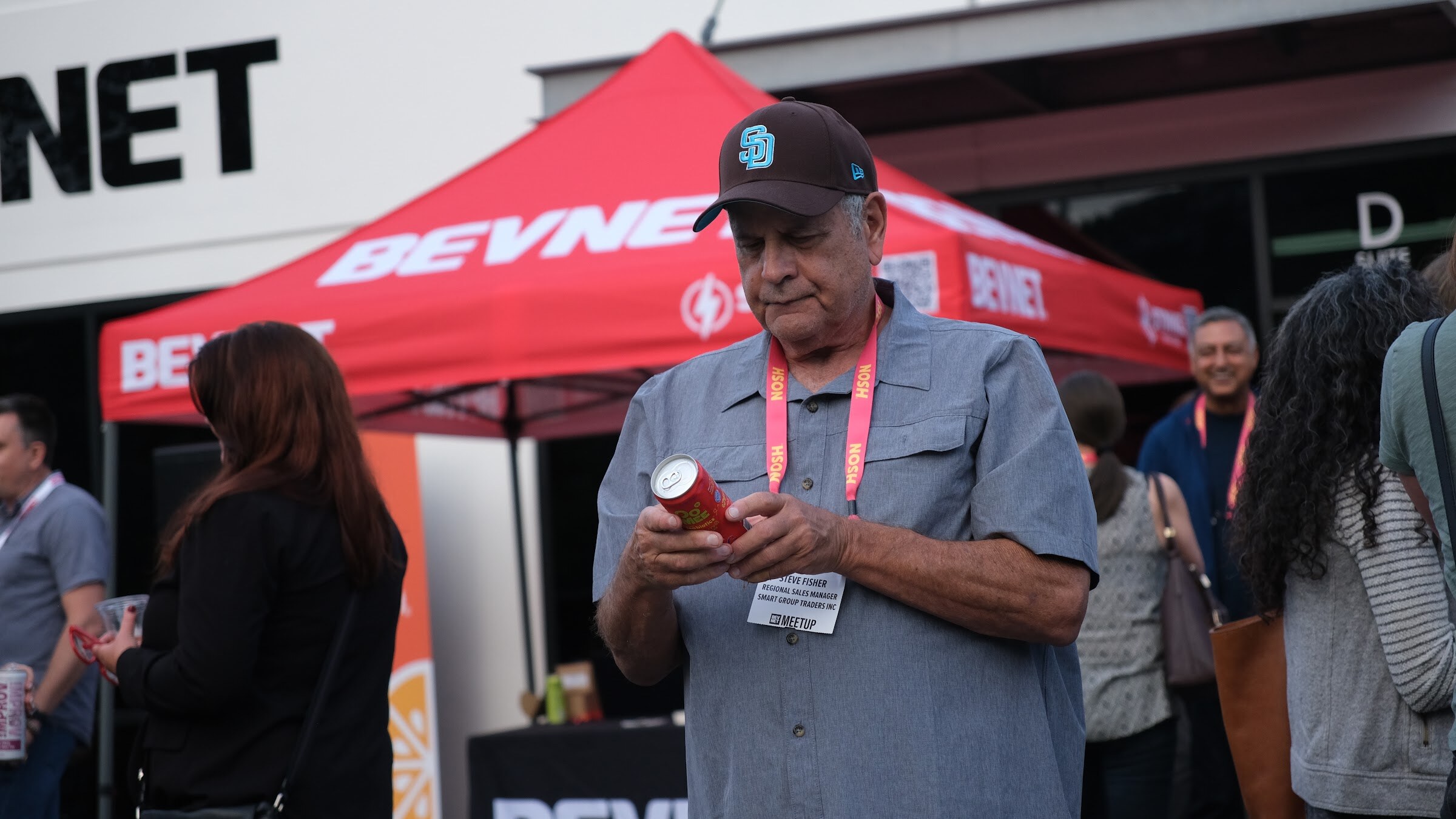 A man in a baseball cap and grey button-up shirt looks at a can he holds in front of a red BEVNET tent.