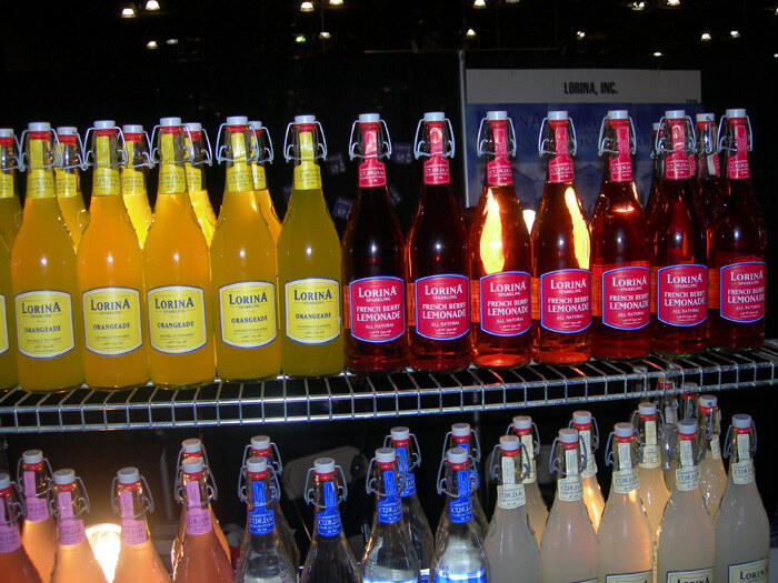 Shelves of swing-top Lorina bottles, including yellow orangeade and red French berry lemonade, with more bottles on a lower shelf and a Lorina, Inc. sign behind.
