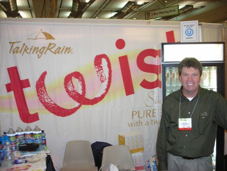 A man in a dark green shirt with a name badge stands in front of a Talking Rain banner with the word twist. Water bottles sit on a table at left and a glass-door cooler stands at right.