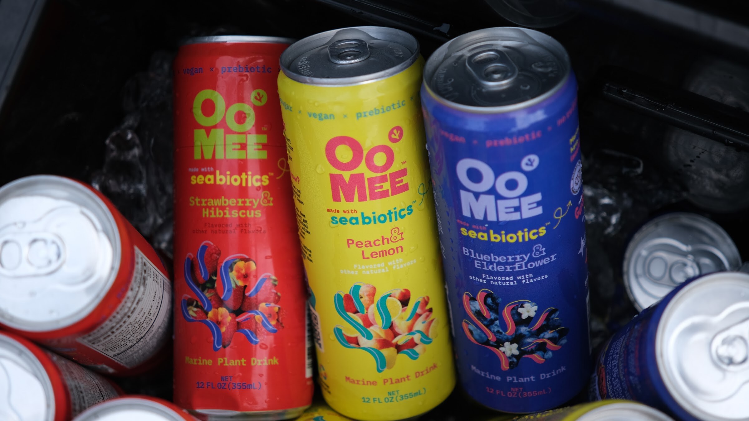 Red, yellow and blue Oo Mee seabiotics cans in strawberry hibiscus, peach lemon and blueberry elderflower flavors.