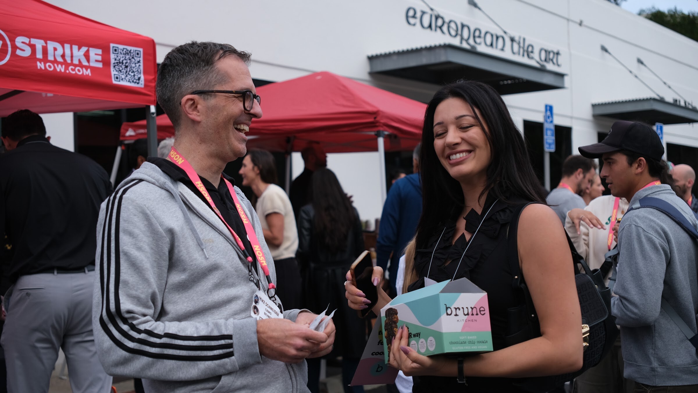 A man in a grey zip hoodie and a woman in black laugh together, she holding a small box labeled brune.