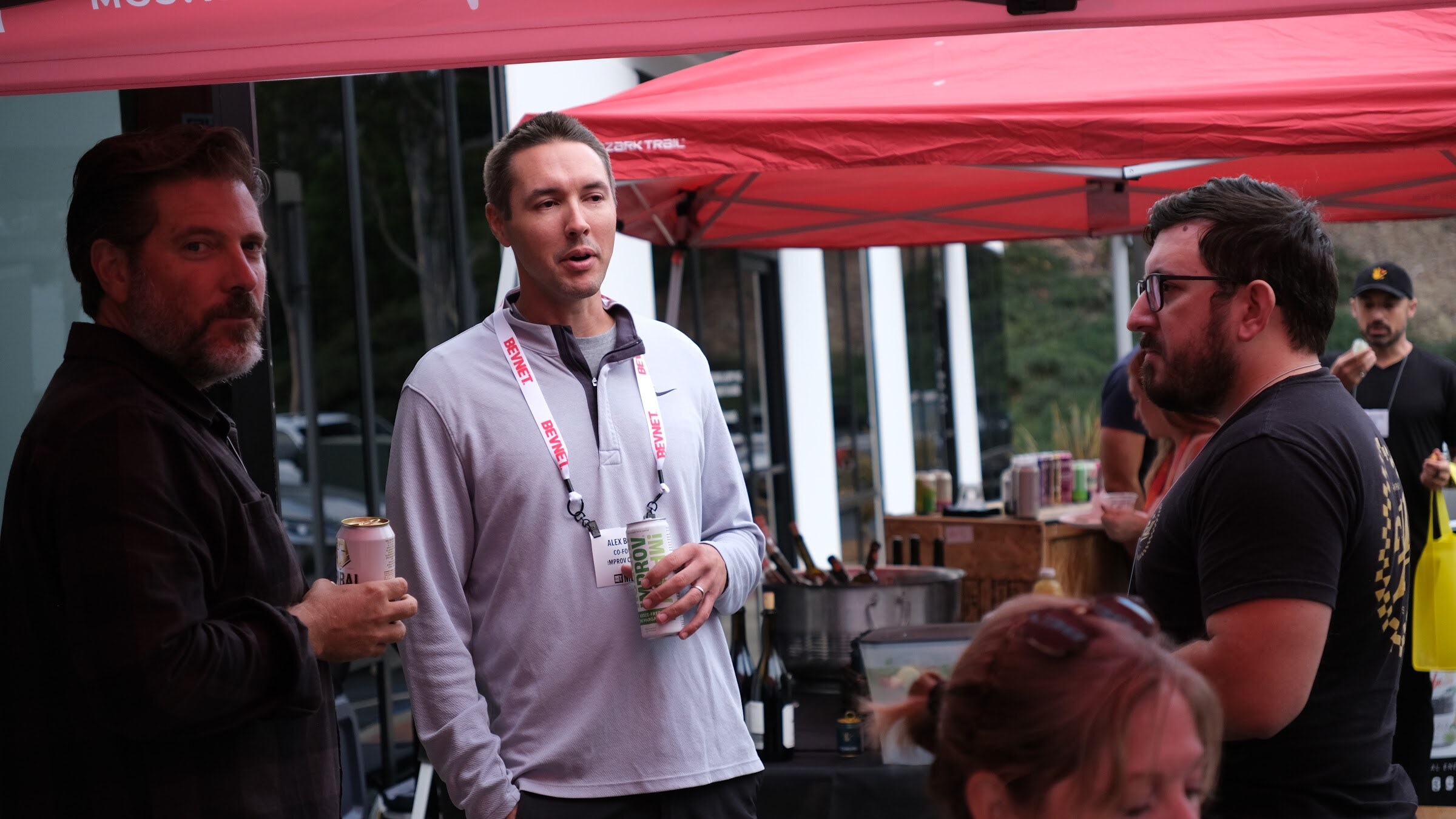 A man in a light grey pullover with a lanyard and a can in hand talks with two other men under red tents.