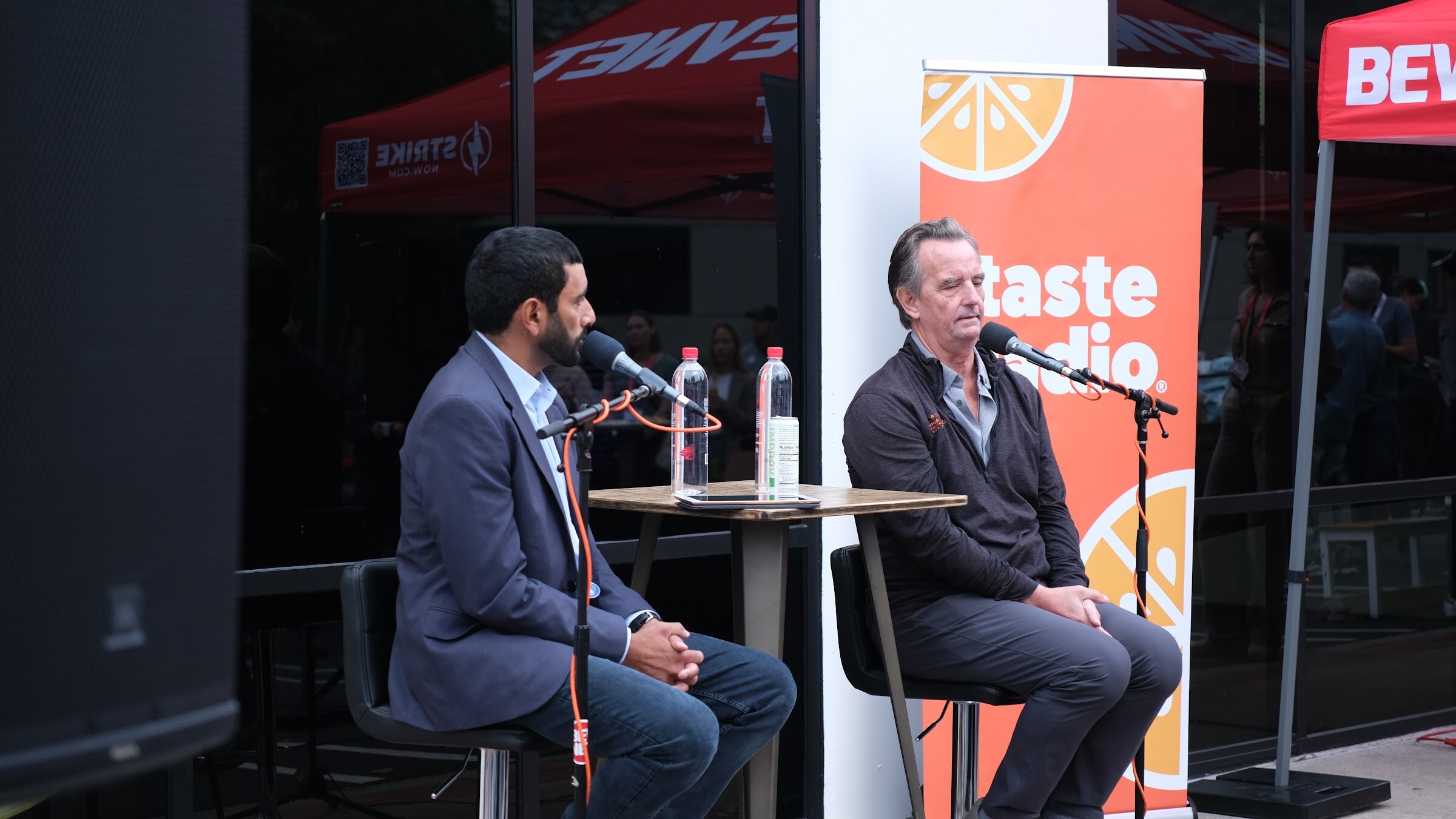 Two men on stools at a small table with microphones and water bottles in front of an orange Taste Radio banner and a red tent.