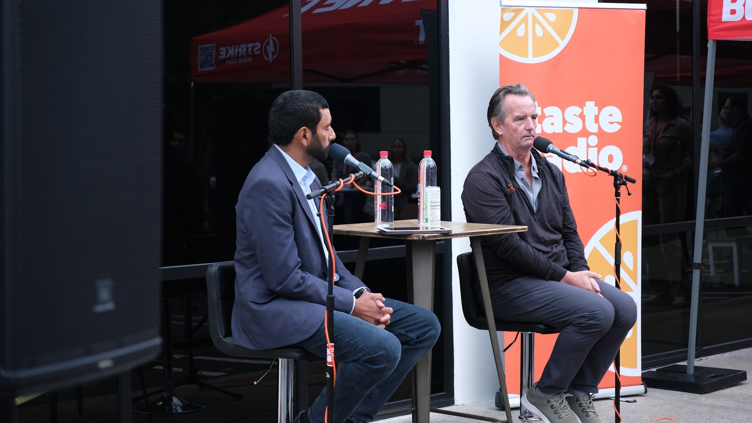 A man in a navy blazer and a man in a dark jacket sit on stools with microphones in front of an orange Taste Radio banner.