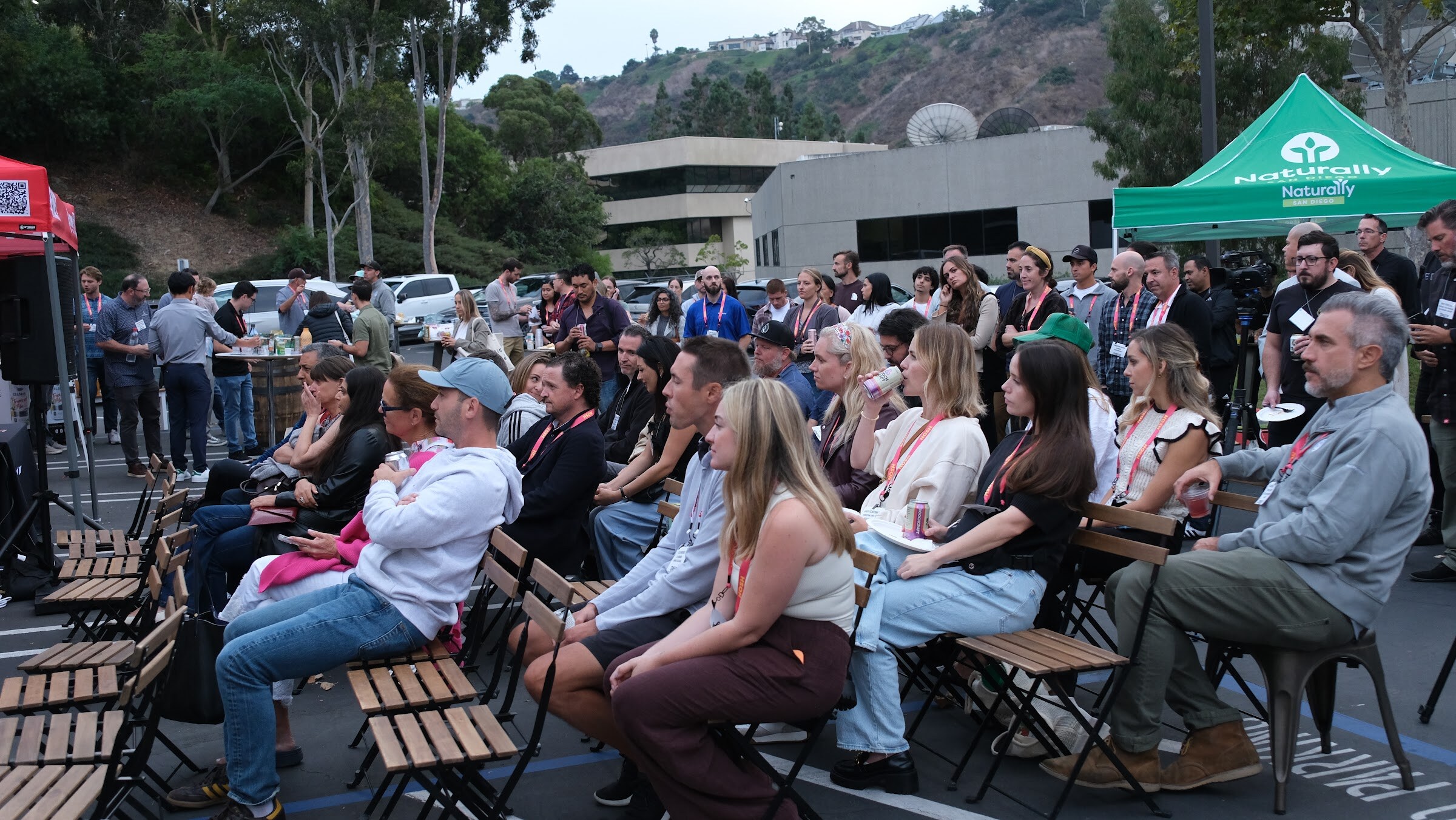Rows of attendees in folding chairs in a parking lot, with a green tent and hills behind them.