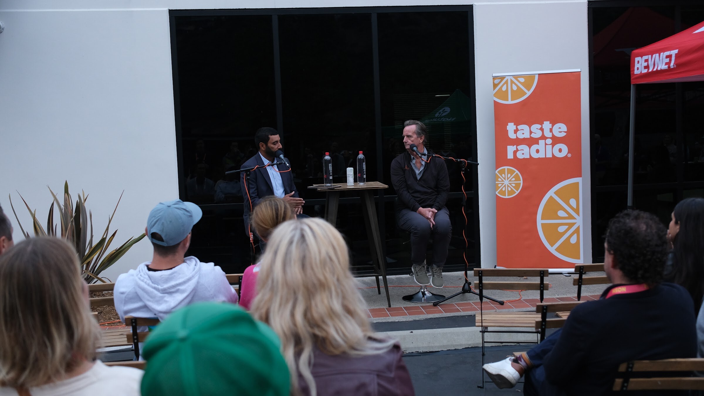 The back of several guests' heads in the foreground facing two speakers seated in front of an orange Taste Radio banner.