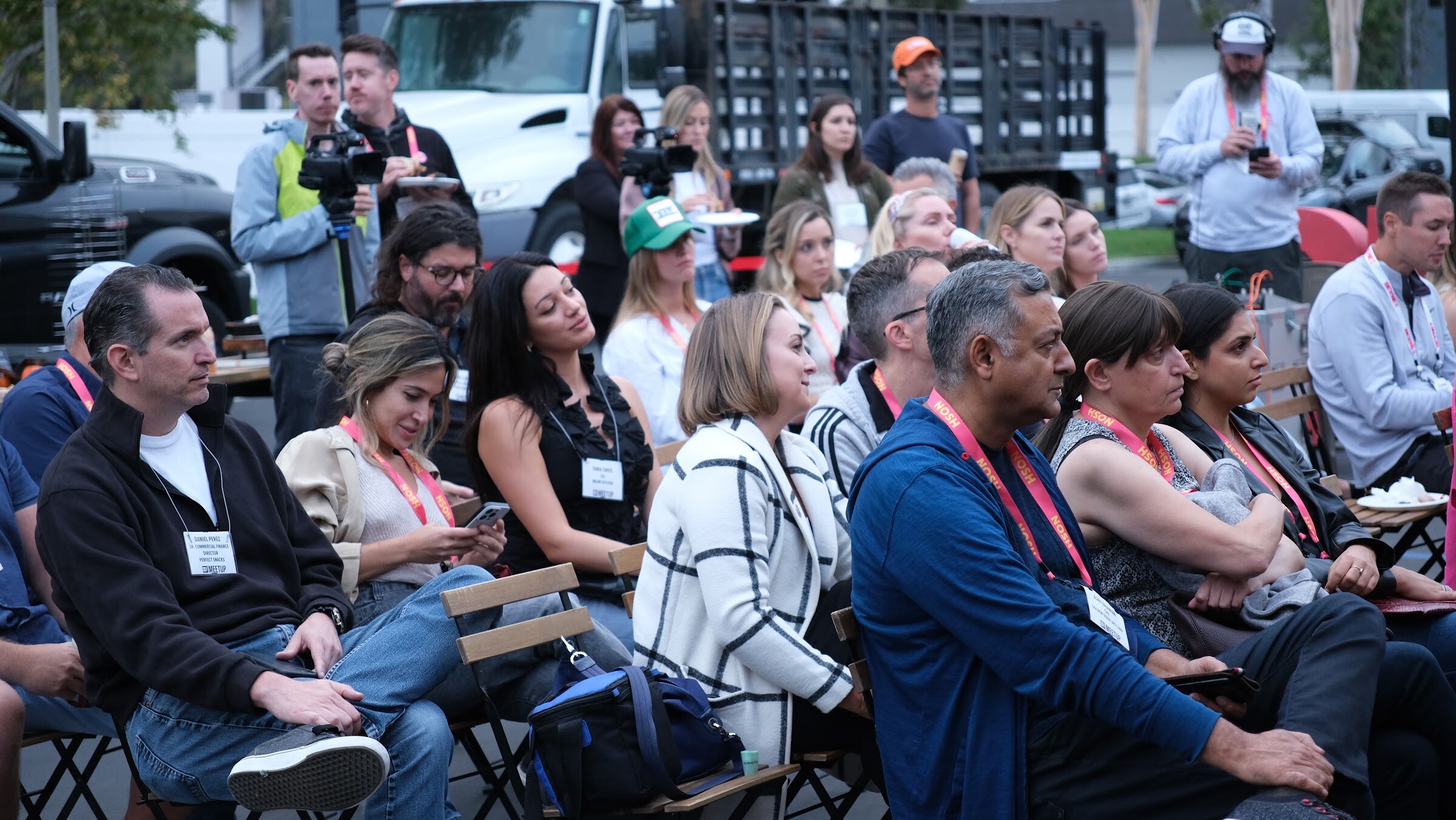 Seated attendees in lanyards, with a camera operator and a truck behind them, watching the interview.