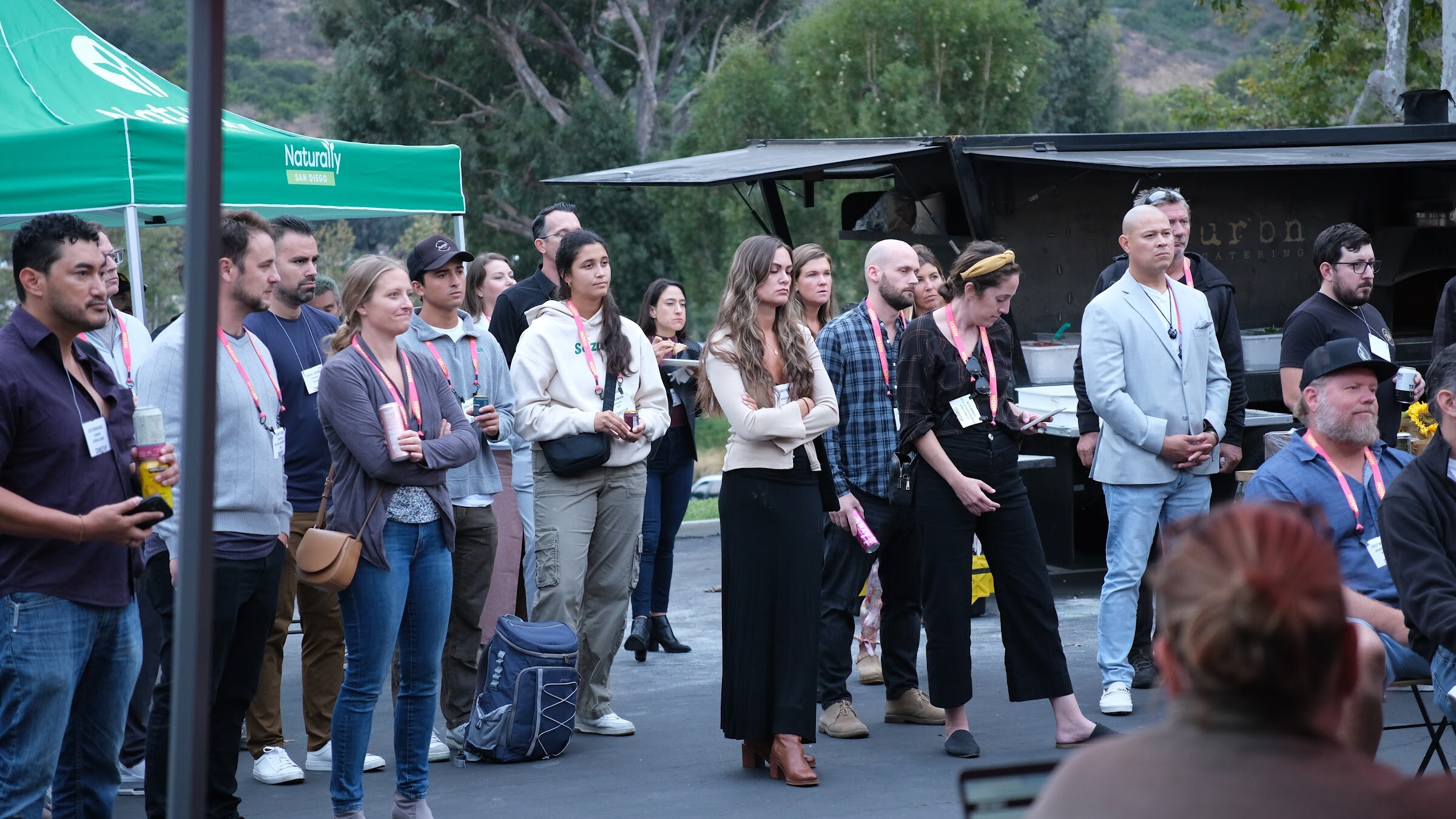 A group of standing attendees in lanyards, with a green tent and a food truck behind them, looking toward the stage.