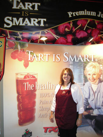 A smiling woman in a maroon apron stands in front of a Tart is Smart banner with cherries and a glass of red juice. A TPG logo appears at the bottom of the display.