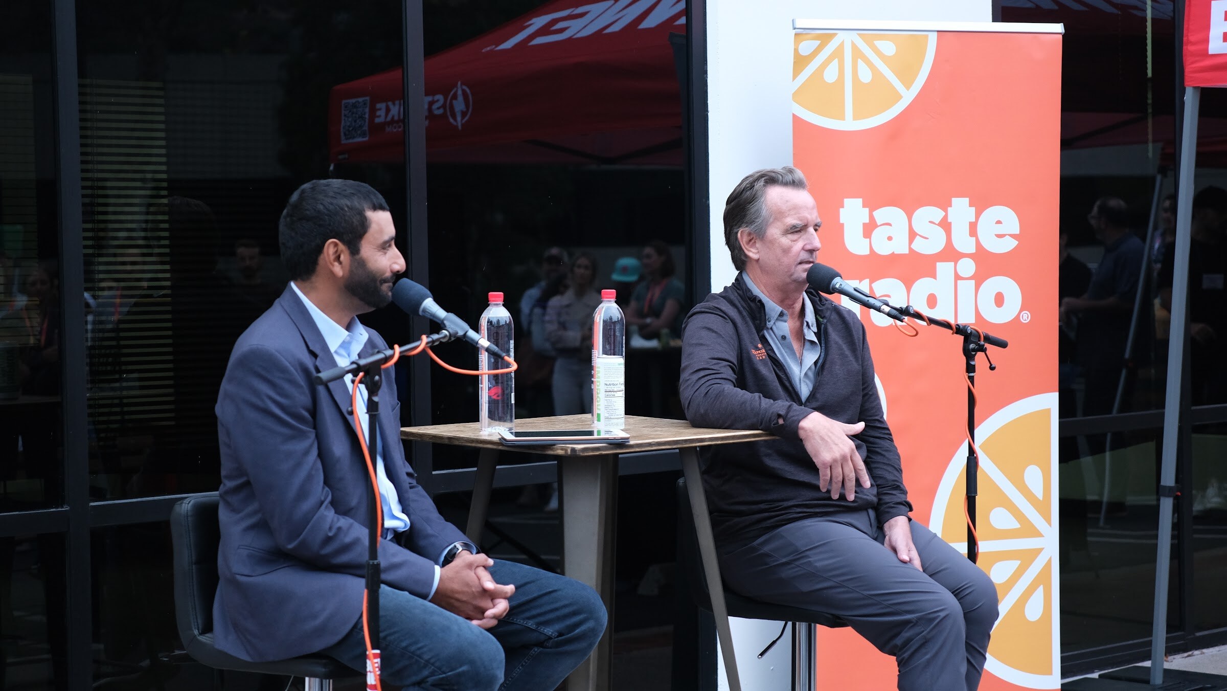 A man in a navy blazer sits with his hands clasped beside a man in a dark jacket speaking into a microphone, with an orange Taste Radio banner behind them.