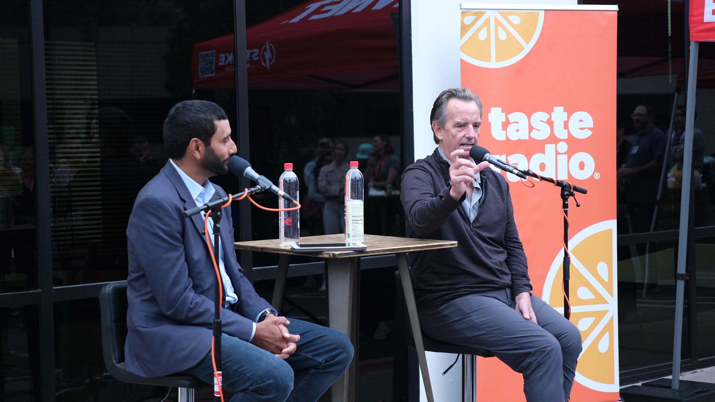A man in a dark jacket gestures with one arm while a man in a navy blazer listens, with an orange Taste Radio banner behind them.
