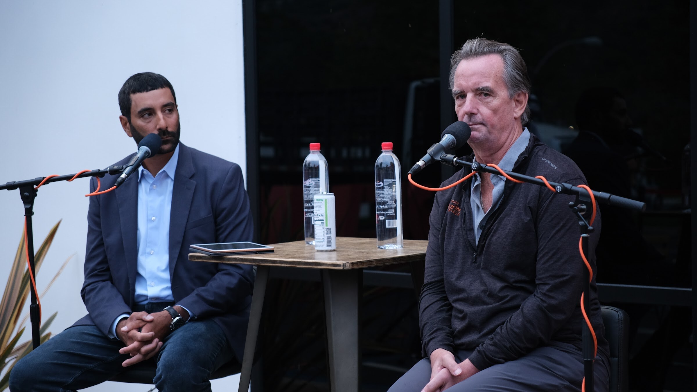 A man in a navy blazer with clasped hands sits beside a man in a dark jacket who is speaking into a microphone.
