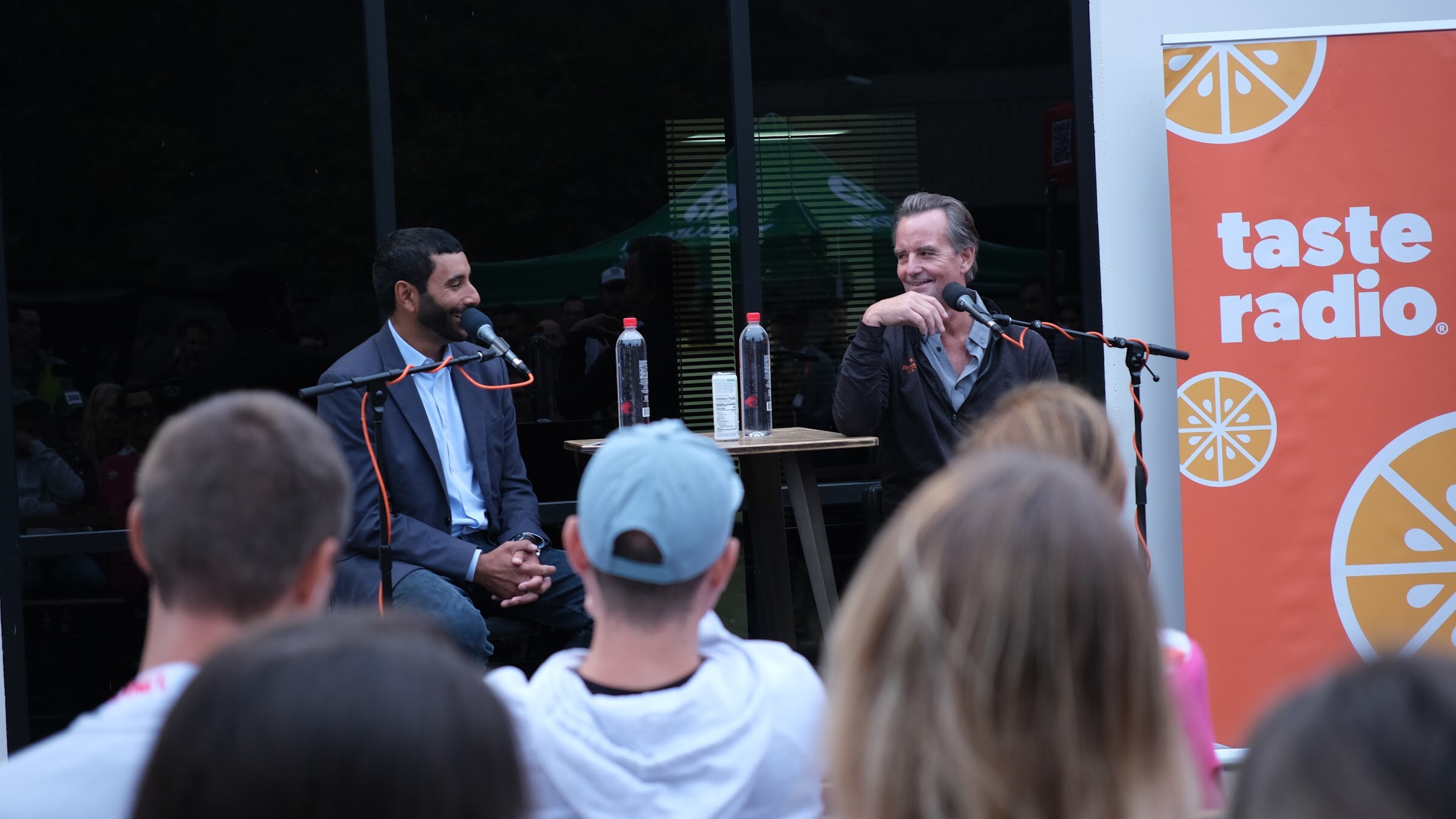 A man in a dark jacket laughs while seated in front of an orange Taste Radio banner, with the backs of audience members in the foreground.