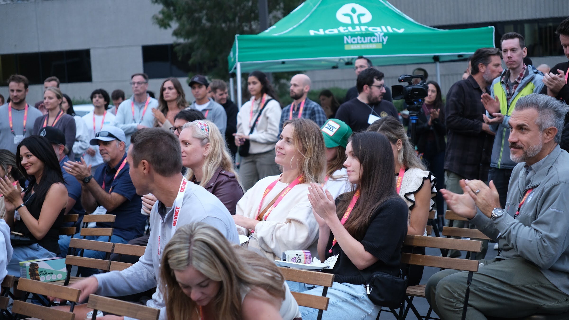 Seated audience members applauding and smiling, with a Naturally tent and camera crew behind them.