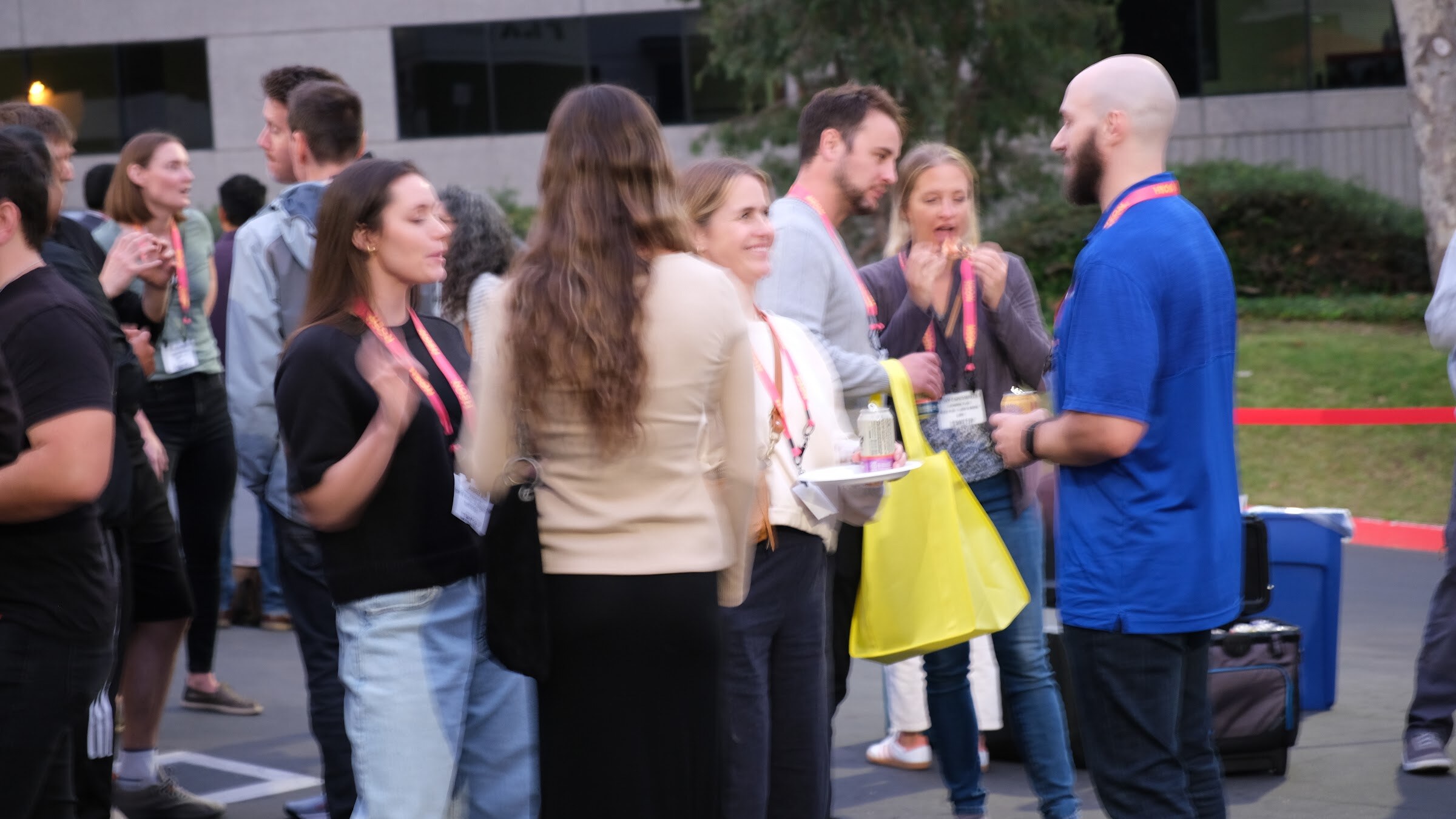 Several attendees talk in a group in a parking lot, one man in a blue polo with a lanyard and a woman holding a yellow bag.