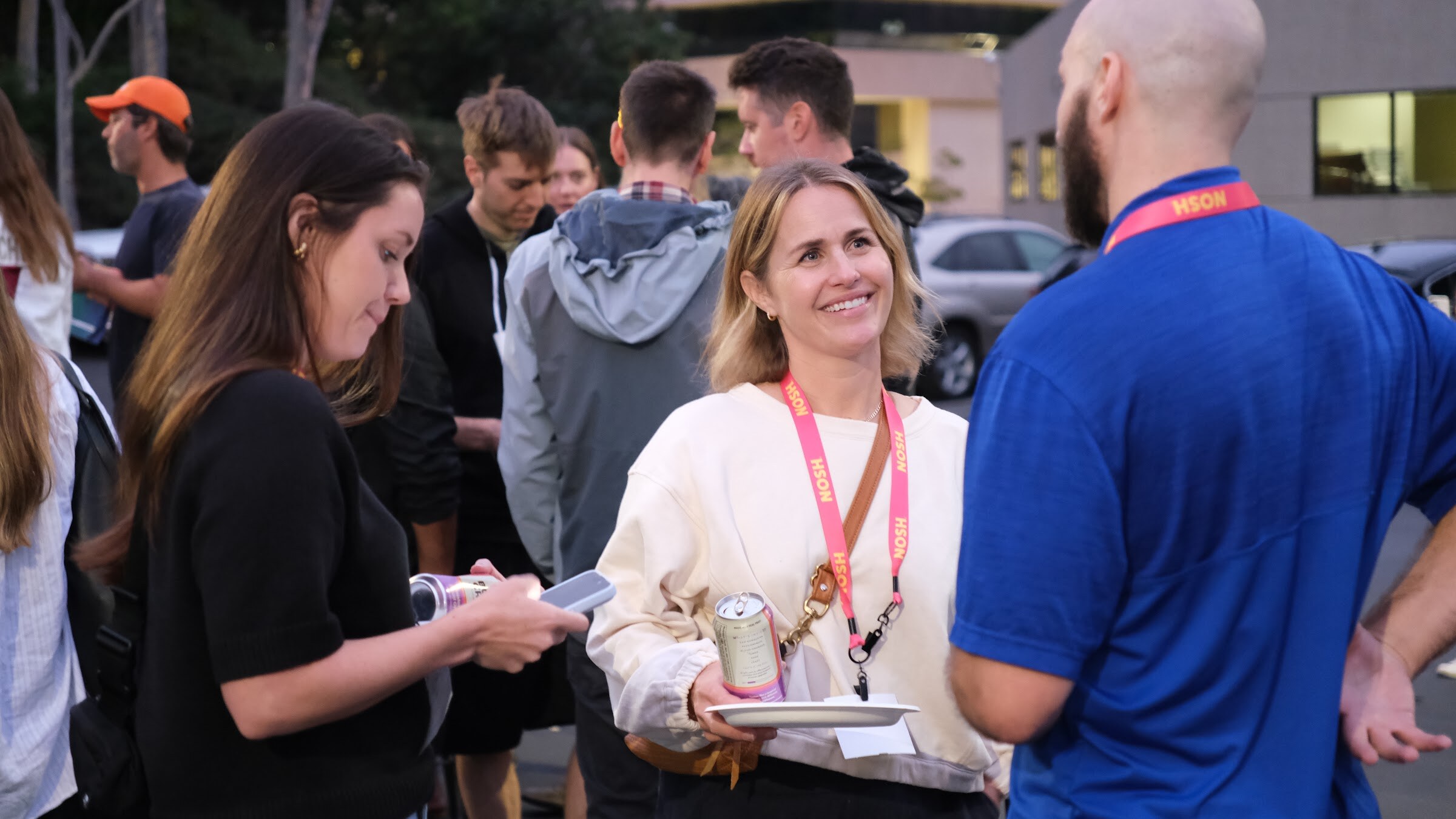 A smiling woman in a cream sweater with a lanyard holds a can and a paper plate while talking with a man in a blue polo.